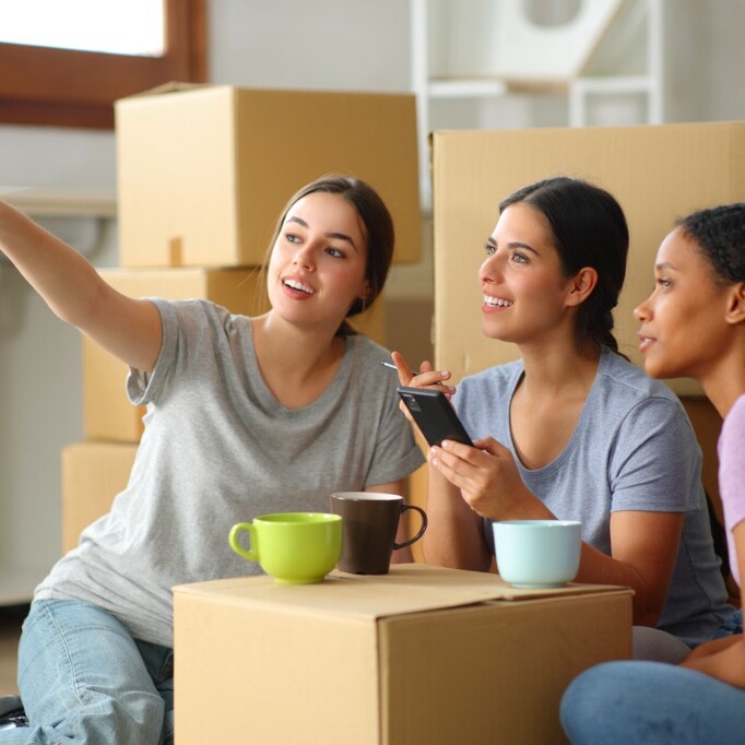 Trois jeunes femmes en colocation assises au sol, entourées de boîtes de déménagement, dans une ambiance conviviale.